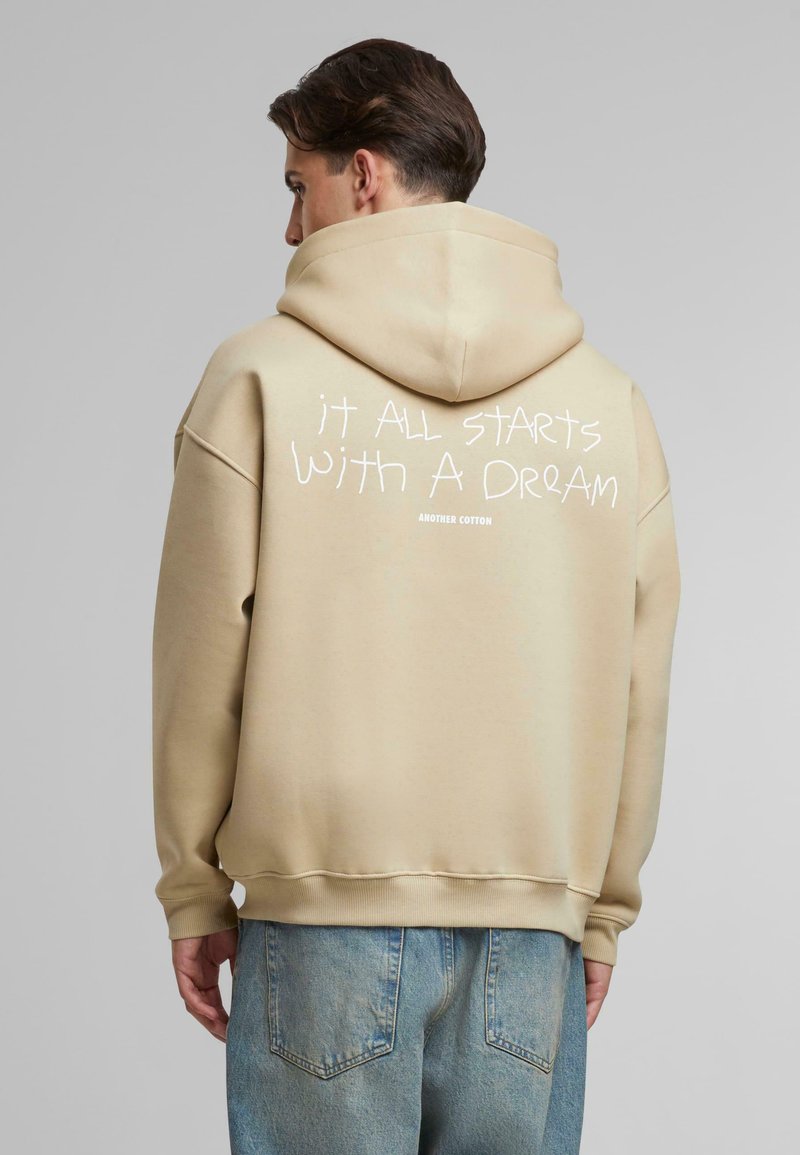 Felpa beige in cotone con vestibilità rilassata, cappuccio oversize e polsini a coste. Presenta una scritta bianca sulla schiena: "tutto inizia con un SOGNO."