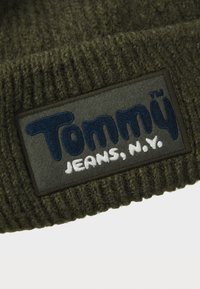 Dark green knitted fabric with a rectangular patch reading "Tommy Jeans, N.Y." in navy and white embroidery.