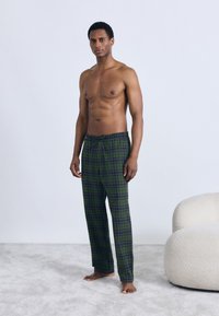 Grønne og marineblå ternede pyjamasbukser med elastisk talje, lavet af blødt stof; båret af en mandlig model, der står på et lyst tæppe.