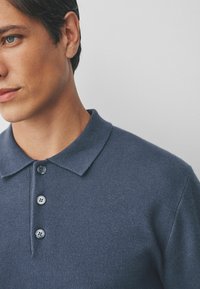 Polo majica tamnoplave boje s mekanom teksturom, kratkim ovratnikom i placketom s tri dugmeta. Pogled izbliza koji ističe detalje tkanine i dizajn ovratnika.