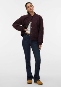 Blouson aviateur matelassé bordeaux avec poignets et col côtelés, associé à un jean évasé en denim foncé et des chaussures marron clair.