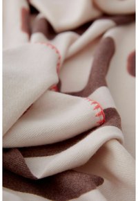 Écharpe en tricot en doux beige avec des motifs irréguliers marron. Présente des coutures texturées en corail vif le long des bords.