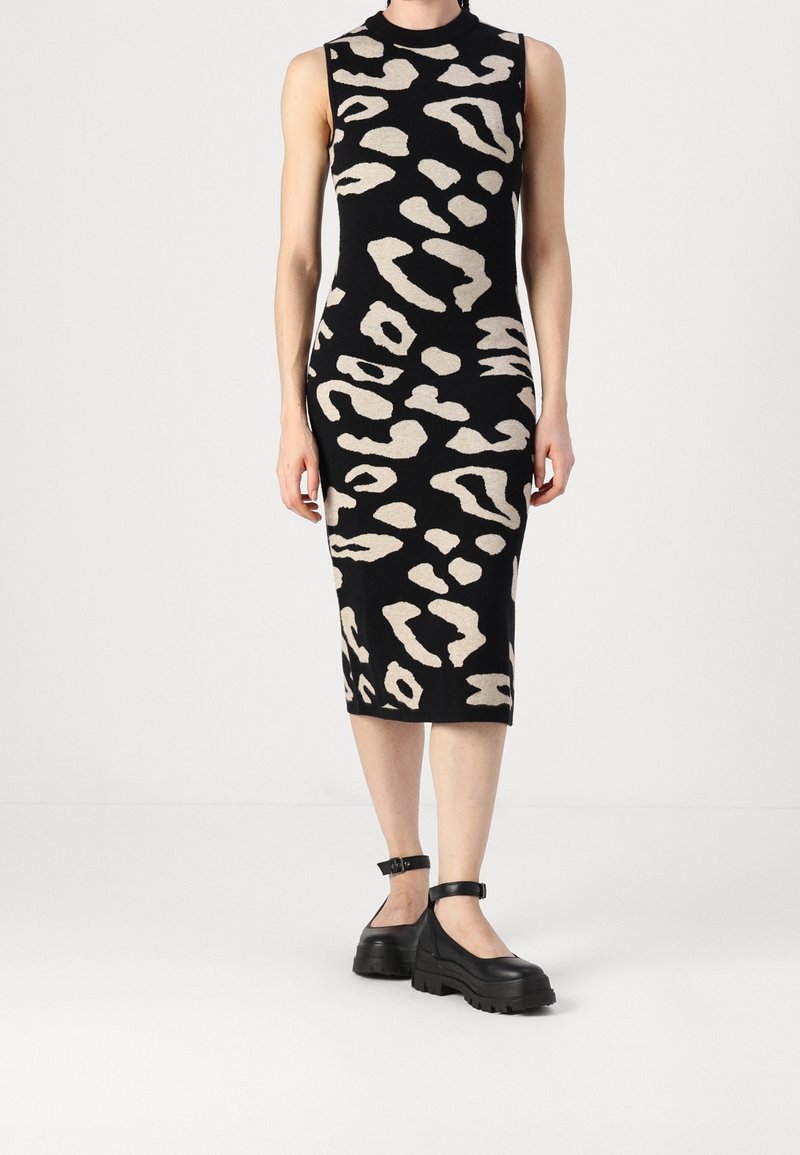 Robe midi noire sans manches avec imprimé animalier abstrait beige, portée avec des chaussures noires épaisses à bride cheville sur fond uni.