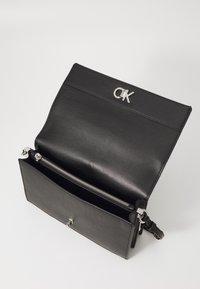 Czarna skórzana torba o gładkim wykończeniu, z srebrnym logo CK, przednią klapką oraz zamkniętym na zamek przegródką wewnątrz.