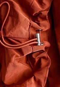 Cordon avec fermoir métallique et œillets enfilés à travers un tissu orange rouille, montrant des détails froncés et cousus.