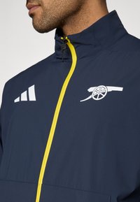 Ναυτικό μπλε αθλητικό σακάκι με κίτρινο φερμουάρ, με λευκά γραφικά λογότυπου κ αι τρεις ρίγες στην αριστερή πλευρά του στήθους. Κατασκευασμένο από ελαφρύ υλικό.