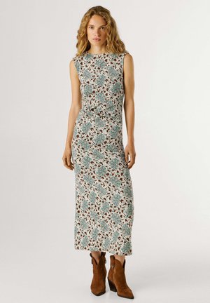 Frau mit welligem blondem Haar, die ein ärmelloses Midikleid mit floralem Muster und braune Wildleder-Stiefeletten trägt, vor einem schlichten Hintergrund stehend.