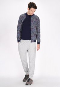 Marine bomberjack met kleurrijke bloemenprint, gecombineerd met lichtgrijze joggingsbroek. Model draagt grijze sneakers met zwarte en rode accenten.