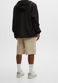 Svart huva jacka, beige shorts, vita strumpor och ljusa skor. Kläderna har en avslappnad passform och minimal design utan synliga mönster.