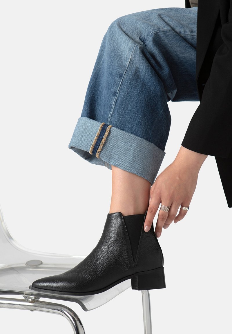 Personne ajustant une botte noire en cuir jusqu'à la cheville en étant assise, portant un jean bleu retroussé et des bagues argentées, reposant le pied sur le siège d'une chaise transparente.