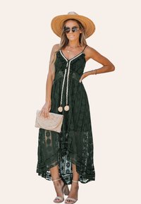 Donkergroene maxi-jurk van kant met een V-hals, bloemenpatronen en zijsplitten, gecombineerd met een geweven clutch en sandaaltjes.