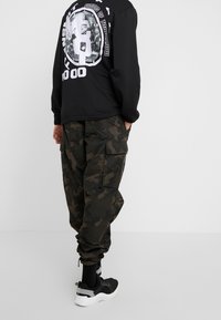 Czarny sweatshirt z białą grafiką na plecach, zestawiony z zielonymi spodniami cargo w kamuflażu, posiadającymi boczne kieszenie i elastyczne mankiety. Czarne sneakersy.