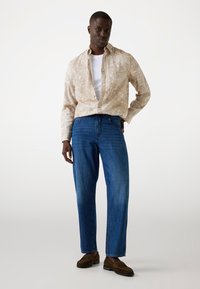 Beige geblokte shirt over een witte t-shirt, gecombineerd met blauwe denim jeans en bruine moccasinschoenen. Casual, relaxte pasvorm.
