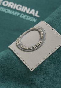 Groene stof met een gestructureerd oppervlak, met een lichtgrijze rechthoekige label die een ronde reliëfontwerp en verhoogde tekst heeft.