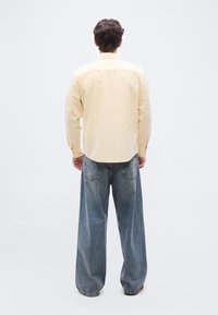 Homme debout, de dos, portant une chemise à manches longues jaune pâle et un jean en denim bleu ample, sur un fond blanc.
