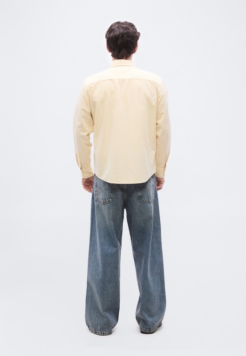 Homme debout, de dos, portant une chemise à manches longues jaune pâle et un jean en denim bleu ample, sur un fond blanc.