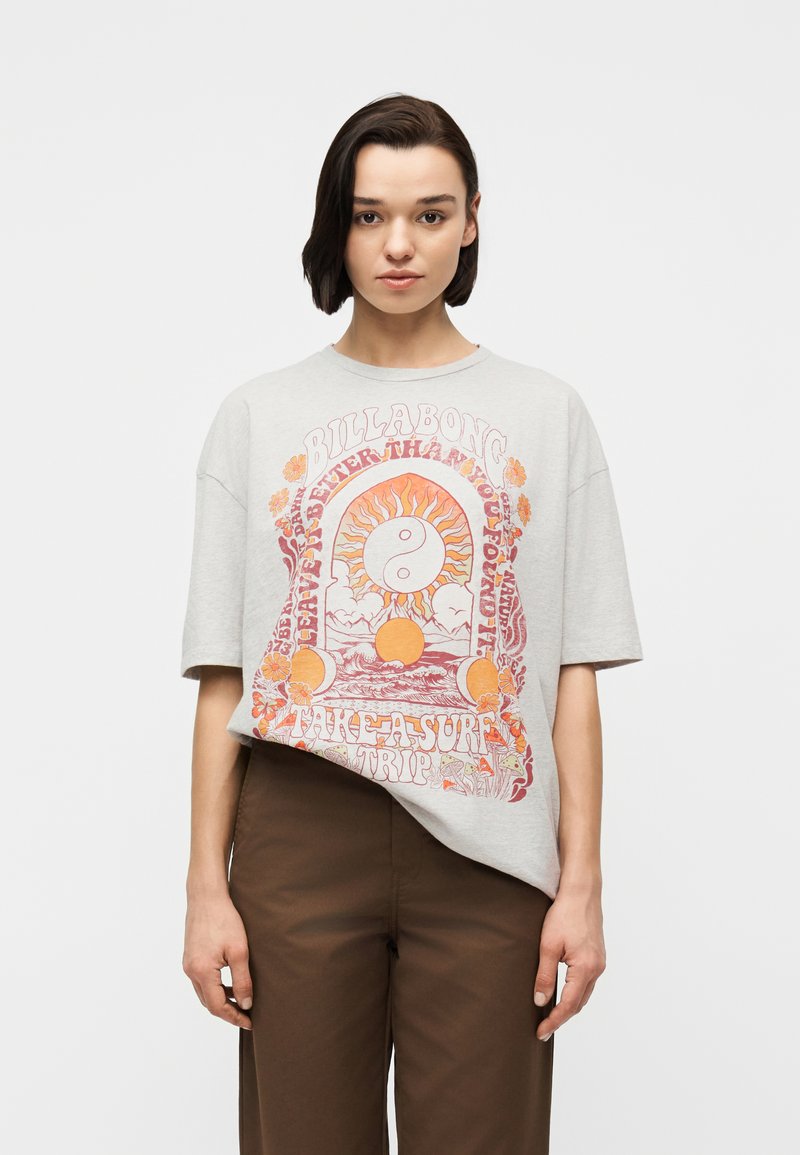 Γυναίκα με κοντά σκούρα μαλλιά που φορεί ένα ανοιχτό γκρι φαρδύ t-shirt με πολύχρωμα γραφικά ήλιου, γιν-γιανγκ και κείμενο, συνδυασμένο με καφέ παντελόνι.