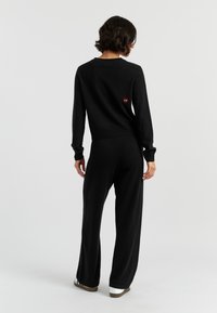 Una persona con capelli ricci indossa un maglione nero e pantaloni abbinati, voltata di spalle. Un piccolo logo rosso è visibile sulla parte posteriore del maglione.
