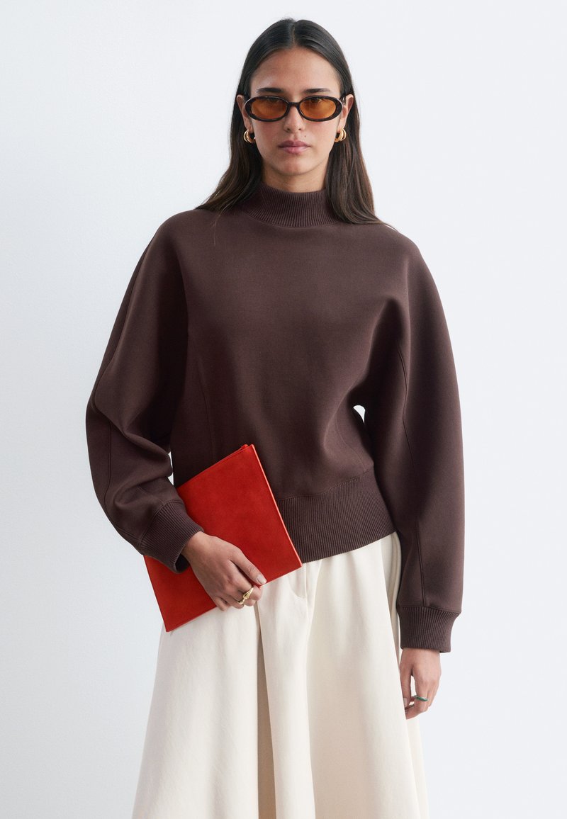 Femme portant un pull à col montant marron, une jupe crème, une pochette orange, des lunettes de soleil ambre et des boucles d'oreilles créoles dorées sur un fond clair uni.