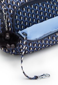 Dragkedjepåse med blå geometrisk mönster, tygmaterial, marinblå botten och svart pom-pom charm fäst med en metallklämma. Slät innervadding.