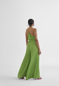 Groene geplooide jumpsuit met dunne kruisbandjes, vloeiende stof en wijde pijpen; gecombineerd met felroze hoge hakken.