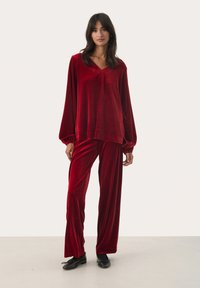 Sametový červený loungewear set obsahující top s dlouhým rukávem a V-výstřihem a široké kalhoty, s nařasenými manžetami a měkkou, hladkou texturou.