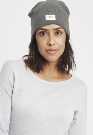 Gray knit beanie with a logo patch, featuring a slouchy design. The subject wears a light gray sweater, highlighting the hat's texture.