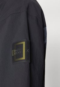 Sort jakke med tekstureret stof, der har et fremtrædende sort og gult logo-patch på venstre ærme. Enkel, funktionel design.