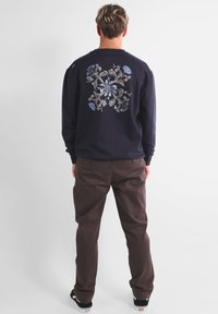 Sweatshirt marine ornée d'une grande broderie florale en bleu et crème dans le dos, associé à un pantalon marron et des baskets noires.