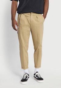 Pantalons en coton beige avec des plis, présentant un détail de chaîne sur le côté. Associés à des baskets noir et blanc et des chaussettes blanches chevillères.