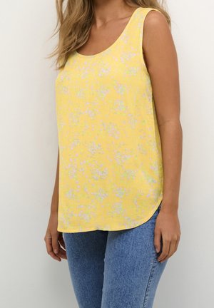 Kvinde iklædt gul ærmeløs top med små hvide blomsterprint kombineret med blå jeans foran en ensfarvet baggrund.