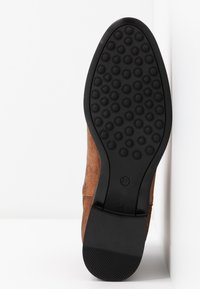 Chaussure en daim marron vue du côté de la semelle, avec une semelle en caoutchouc noir comportant des motifs de bande de roulement circulaires et une adhérence texturée au talon.