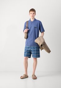 Blå polo med bröstficka, jeansshorts med ett vitt rutmönster, beige jacka och bruna sandaler. Avslappnad outfit utan accessoarer.