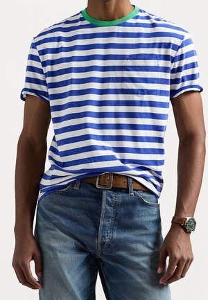 Mężczyzna w niebiesko-białą w paski koszulce z zielonym kołnierzykiem, niebieskich dżinsach, brązowym skórzanym pasku oraz zegarku na nadgarstku z brązowym paskiem i zieloną tarczą.