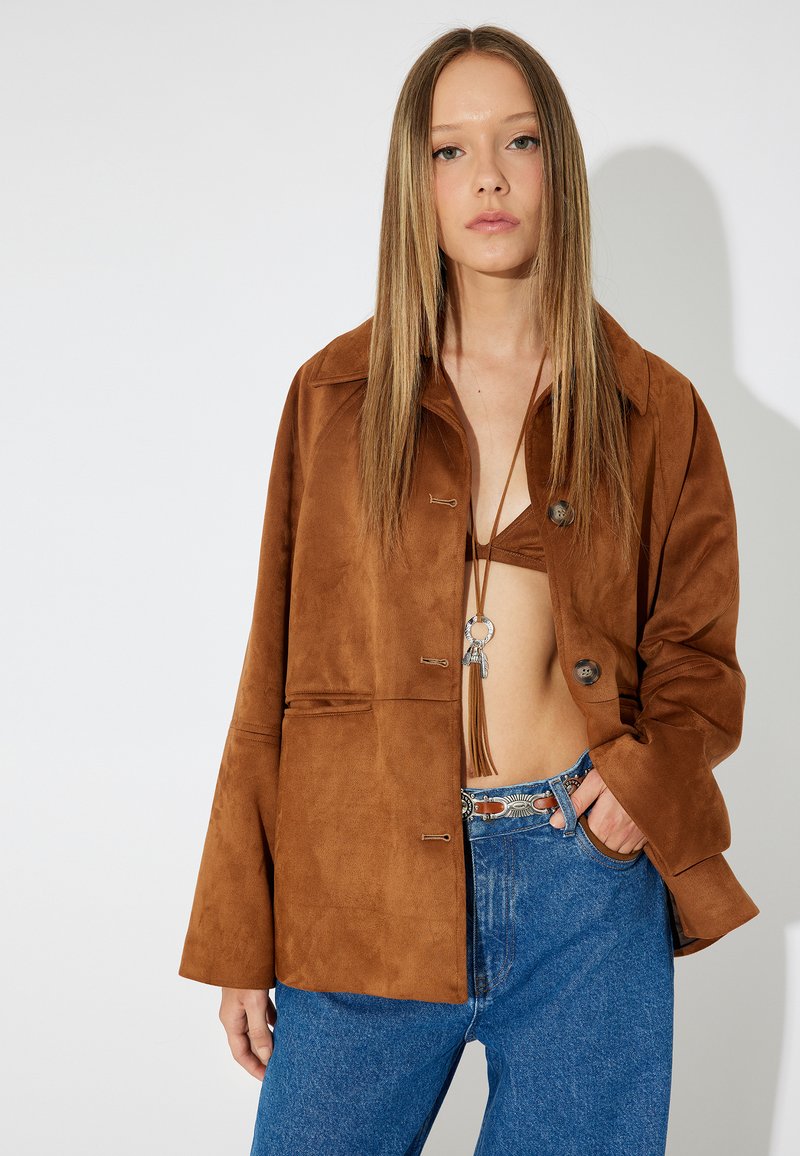 Braune Wildlederjacke mit Kragen, drei Knöpfen und lockerer Passform. Getragen über einem Bralette, kombiniert mit blauen Jeans und einem dekorativen Gürtel.