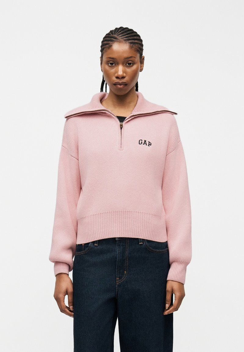 Femme aux cheveux tressés portant un pull à moitié zippé rose pâle avec le logo "GAP" et un jean bleu foncé, sur un fond clair uni.