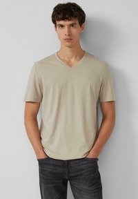 Beżowy t-shirt z dekoltem w serek, wykonany z miękkiego materiału, z krótkimi rękawami i subtelnym logo na piersi. Noszony z ciemnymi dżinsami.