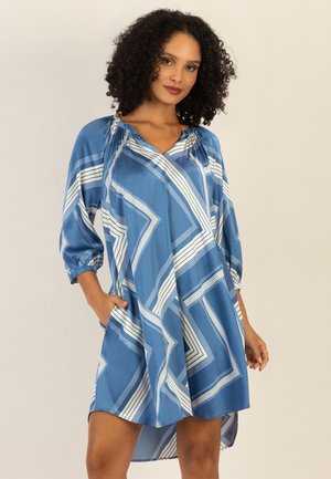 Frau mit lockigem Haar, die ein knielanges Kleid mit blau-weißem geometrischem Muster und dreiviertellangen Ärmeln trägt, steht mit einer Hand in der Tasche.