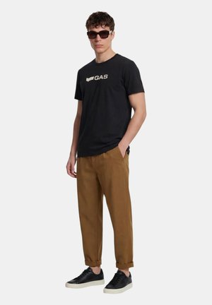 Giovane uomo con occhiali da sole neri, t-shirt nera con logo "GAS", pantaloni marroni risvoltati e sneaker nere con suole bianche.