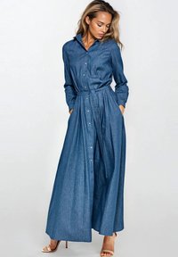 Kobieta z długimi blond włosami, ubrana w długą niebieską dżinsową sukienkę maxi z długim rękawem, z zapięciem na guziki i przewiązaną w talii, oraz w beżowe sandały na obcasie.