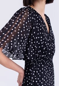 Czarna sukienka w białe kropki z krótkimi, przezroczystymi rękawami, głębokim dekoltem w kształcie V i marszczoną talią. Materiał wydaje się lekki i teksturowany.