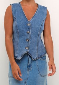 Ärmellose blaue Jeansweste mit vier Knöpfen, getragen über einem passenden blauen Jeansrock, die Arme entspannt an den Seiten.