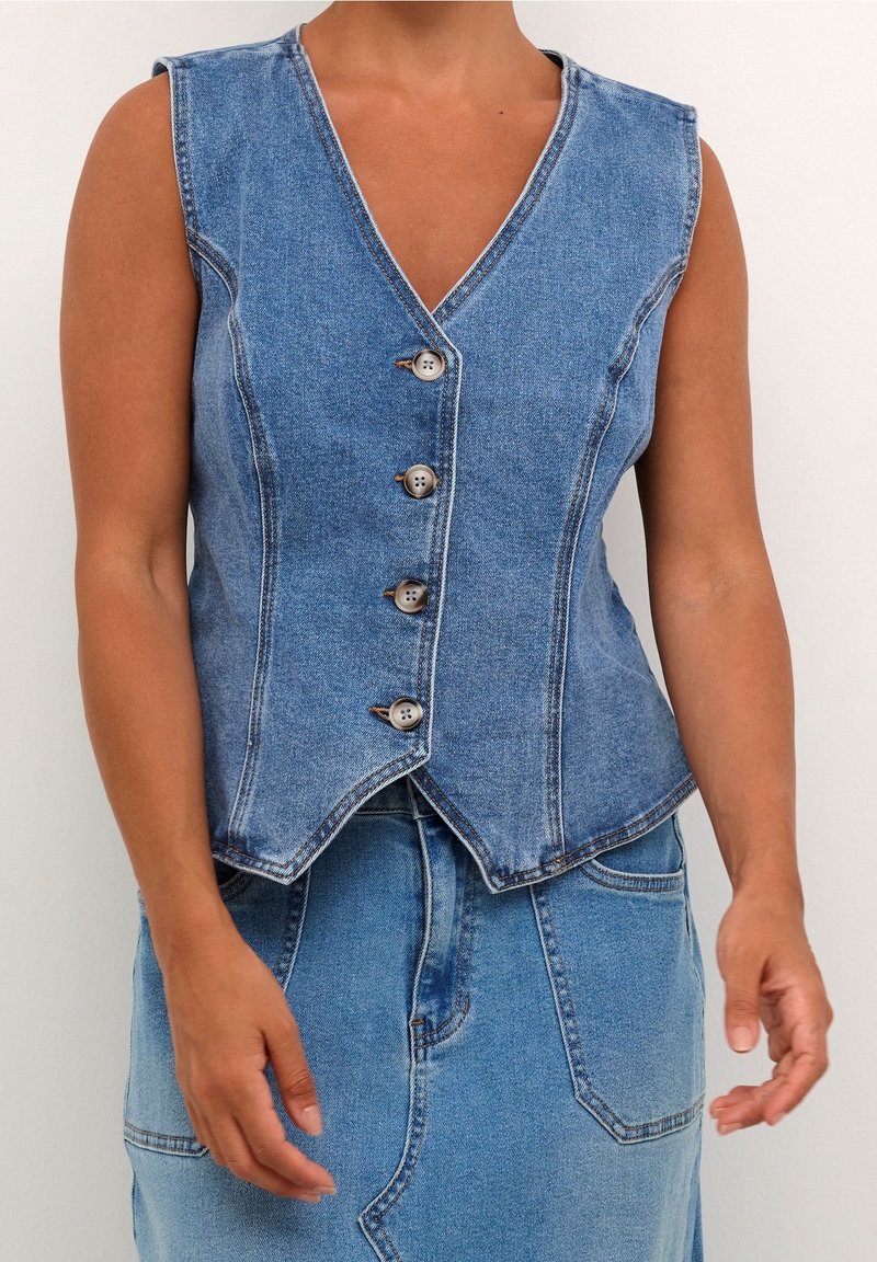 Ärmellose blaue Jeansweste mit vier Knöpfen, getragen über einem passenden blauen Jeansrock, die Arme entspannt an den Seiten.
