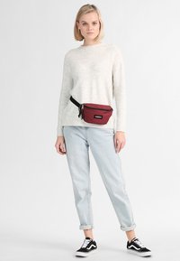 Riñonera roja con un bolsillo con cremallera, llevada en la cintura. Modelo con un suéter claro y jeans azules claros enrollados, con zapatillas negras.