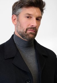 Zwarte wollen overjas met een geknipt kraag, enkele knoopsluiting en een grijze coltrui. Kenmerkt zich door gladde texturen en een getailleerde pasvorm.