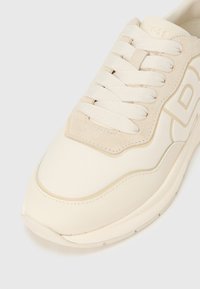 Šviesiai smėlio spalvos sportbačiai su lygiu viršumi, zomšos detalėmis ir plokščiomis baltomis raištelėmis. Batai turi apvalią nosį ir subtilias siuvimo detales.