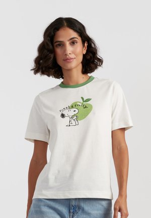Frau mit lockigem dunklem Haar, die ein weißes T-Shirt mit Snoopy trägt, der einen grünen Apfel hält, mit dem Schriftzug "Frisch & Fruchtig", kombiniert mit blauen Jeans.