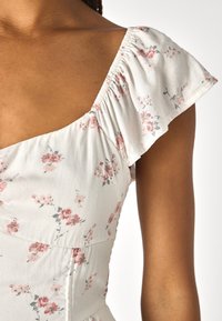 Vit blommig klänning med rosa och gröna mönster, med en åtsittande livstycke och volangärmar. Slät tyg med subtil textur.