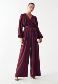 Burgundijska svila kombinezon z globokim V-izrezom, dolgimi rokavi in pasom. Ima široke noge in mehko, prosto padajočo tkanino. Zlata torbica kot dodatek.