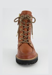 Bottine en cuir marron à lacets avec un bout rond, semelle épaisse noire, accents en métal doré et design de lacets tressés. Dispose de zips latéraux.